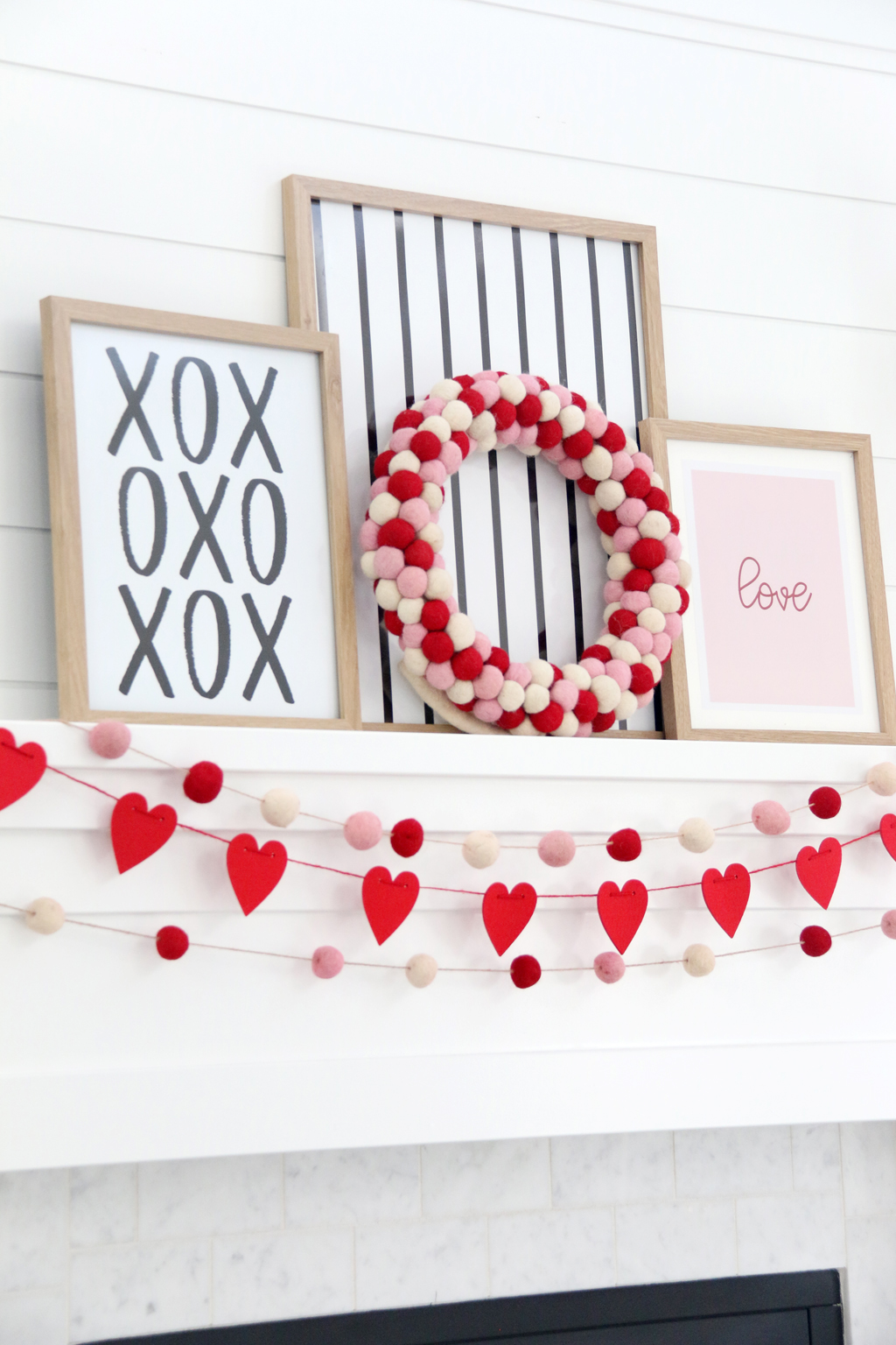 White mantle with red heart banner and pink, red, and white pom pom banner. Three pictures in frames one with XOXO in black and white, one with black and white stripes, and one with Pink background and the word "Love" in red.