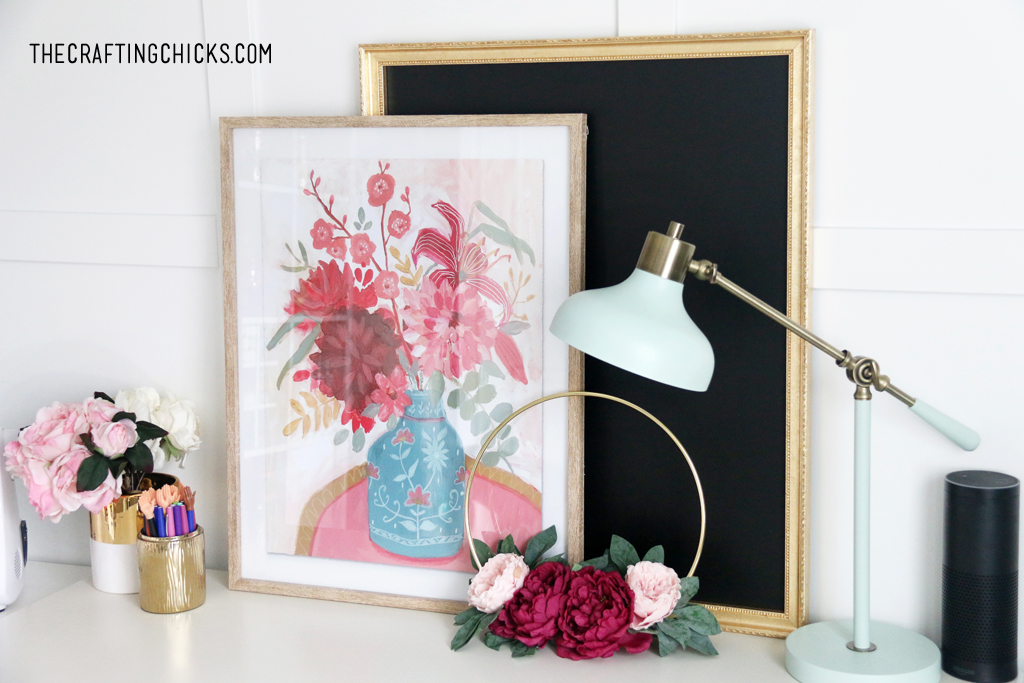 Floral picture in a frame in front of a black chalkboard with gold frame. Floral wreath and blue lamp.