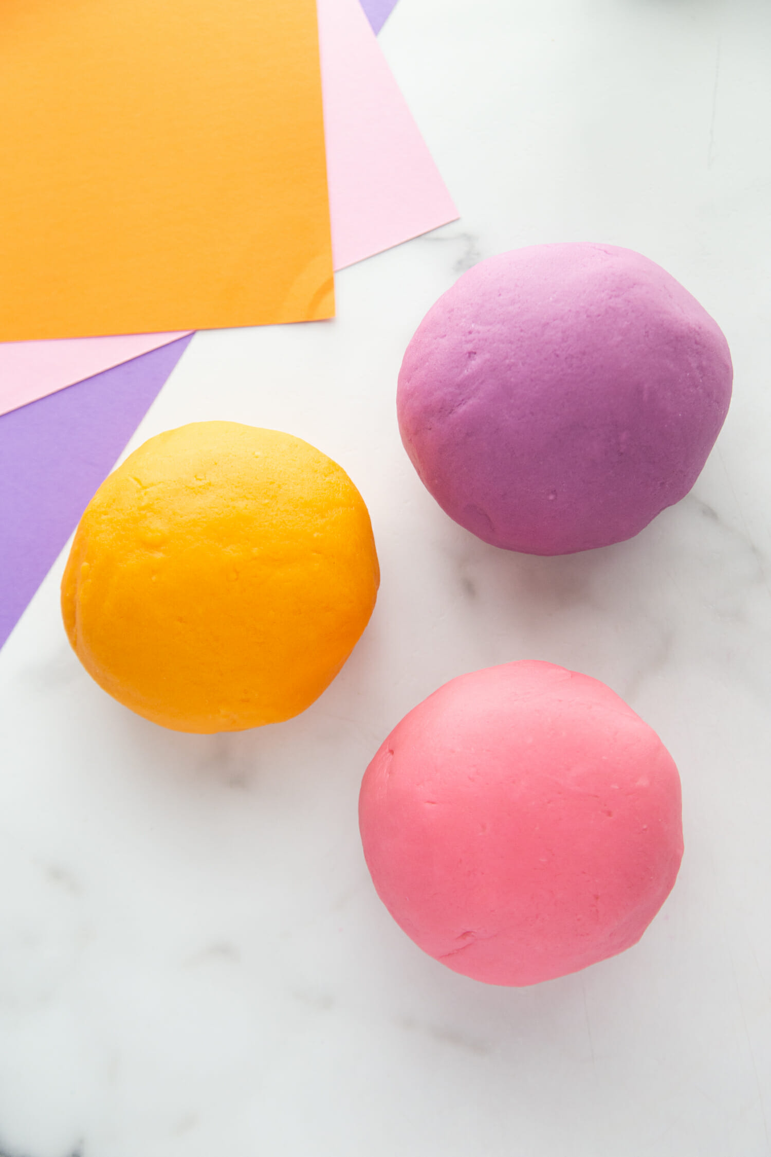 Orange, purple, and pink playdough balls on a white marble countertop.