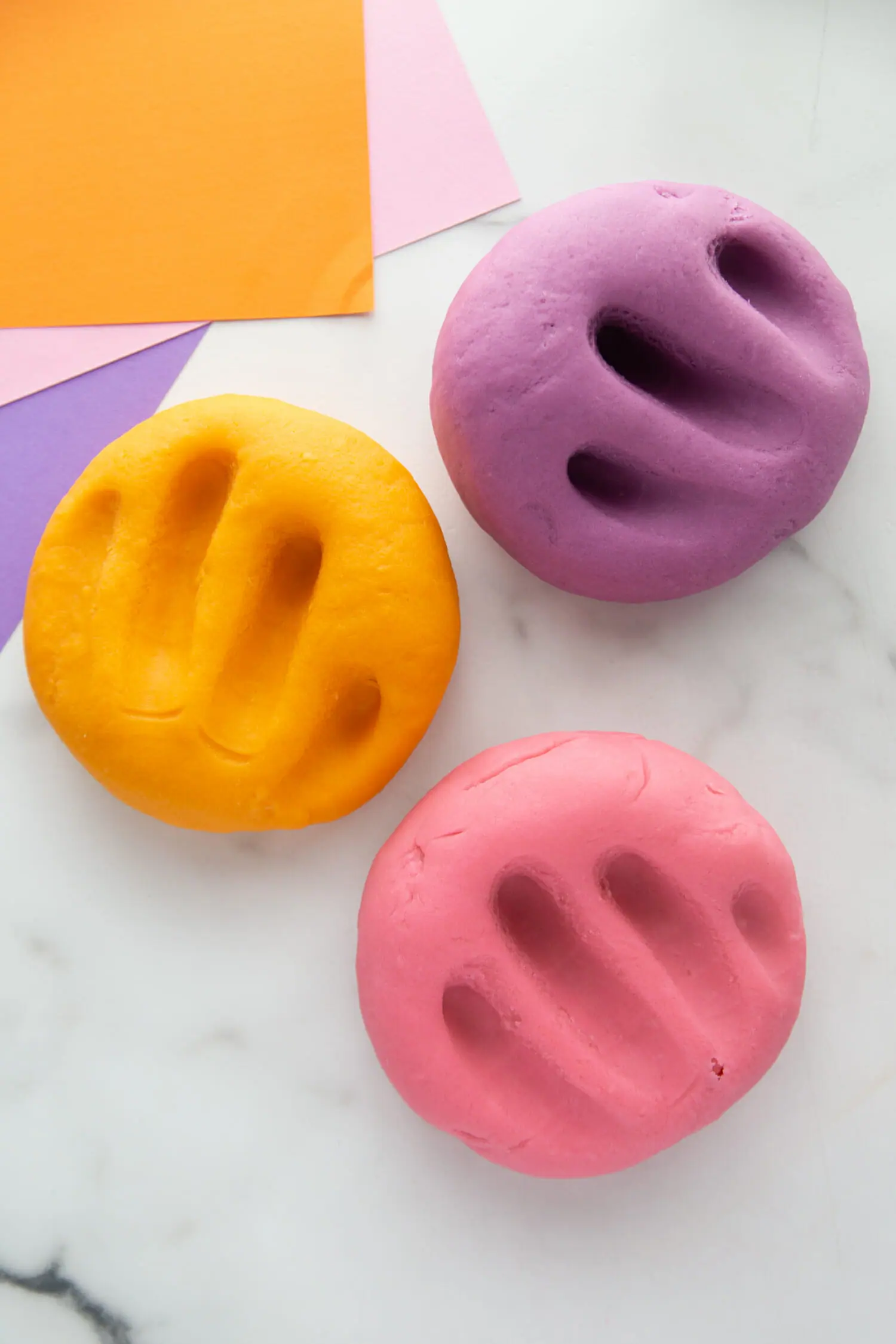 Orange, pink, and purple playdough balls on a countertop with fingers pressed into them. 