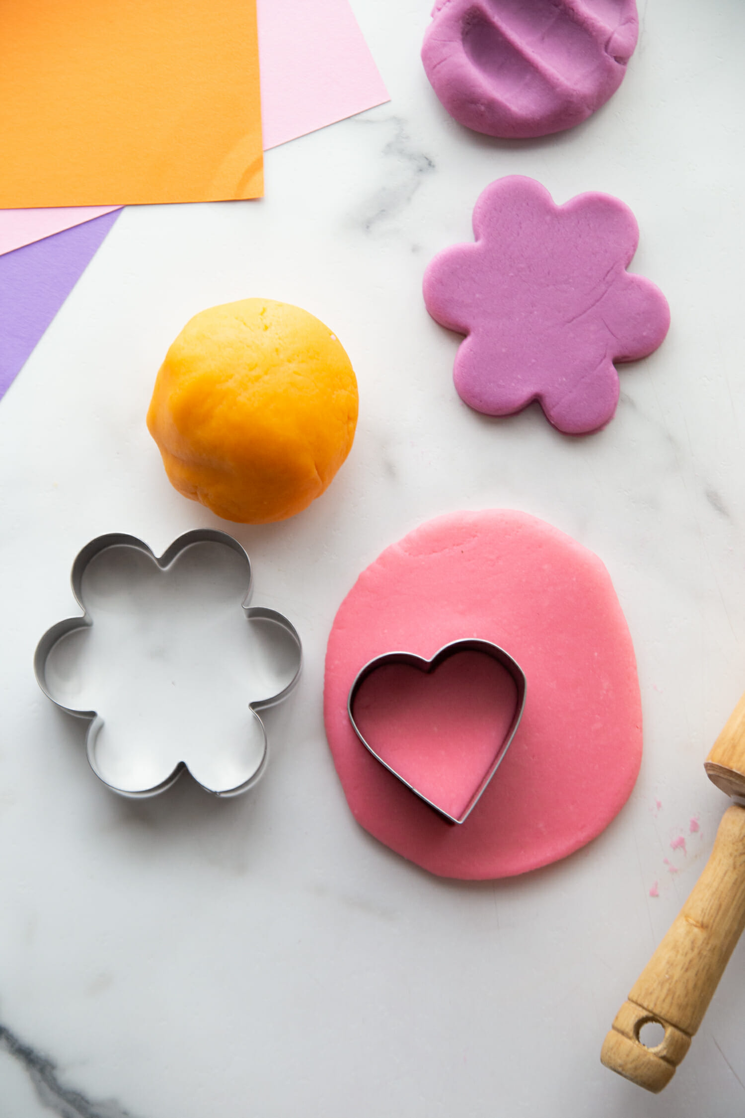 Orange playdough ball next to a purple playdough flower, and pink playdough flatended out with a heart shapped coookie cutter on top.