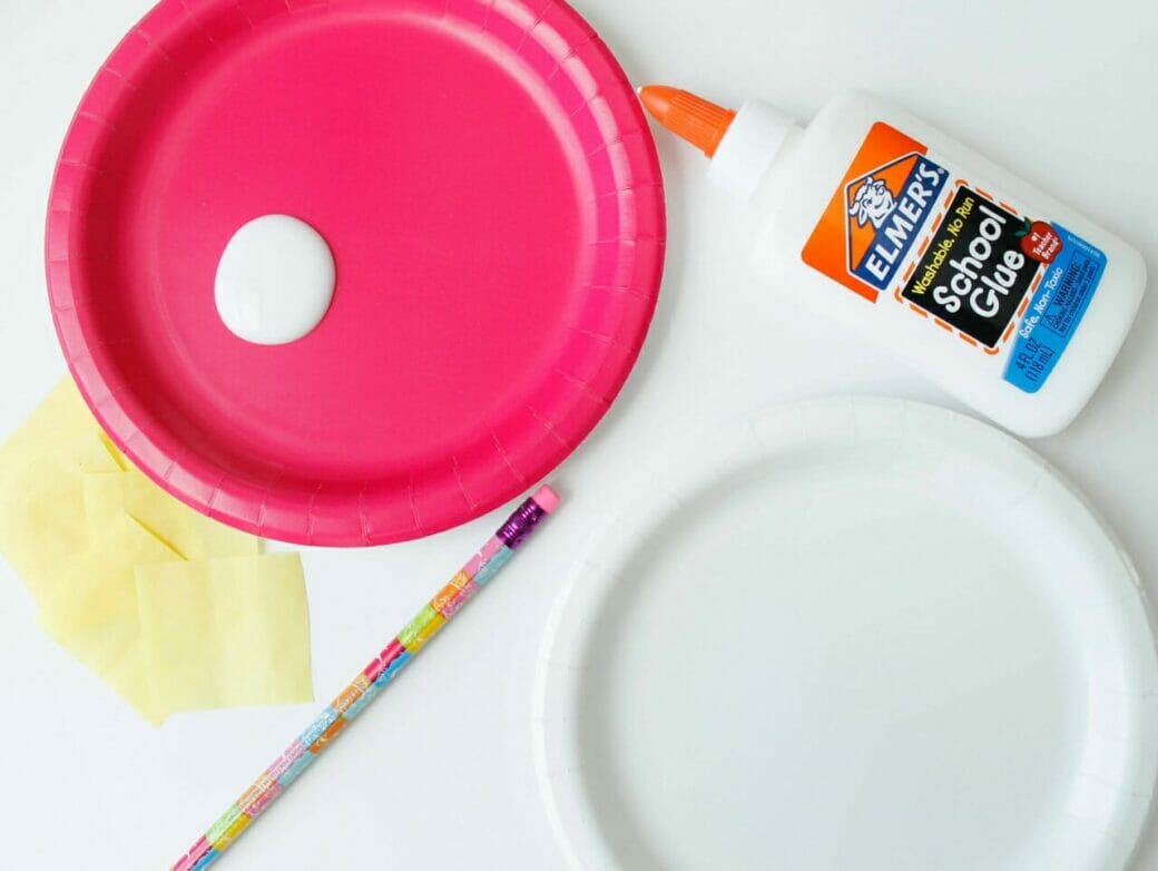 Pink paper plate with white school glue on it, white school glue bottle on the side, yellow tissue paper squares on the side, a pencil, and white paper dessert plate.