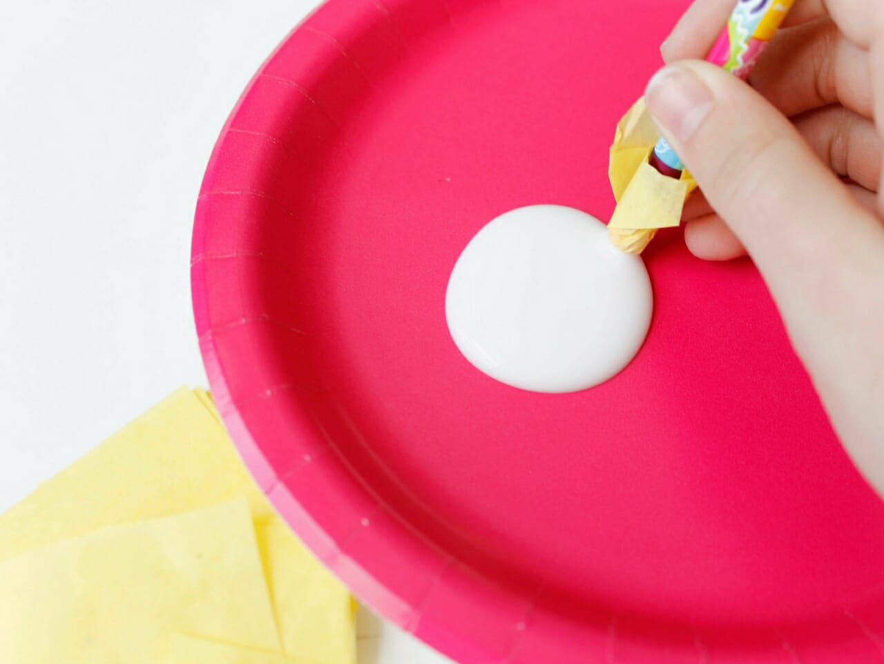 Pink paper plate with some white school glue on it. Yellow tissue paper squares on the side. Hand holding a pencil with tissue paper on it dipping tip into the glue.