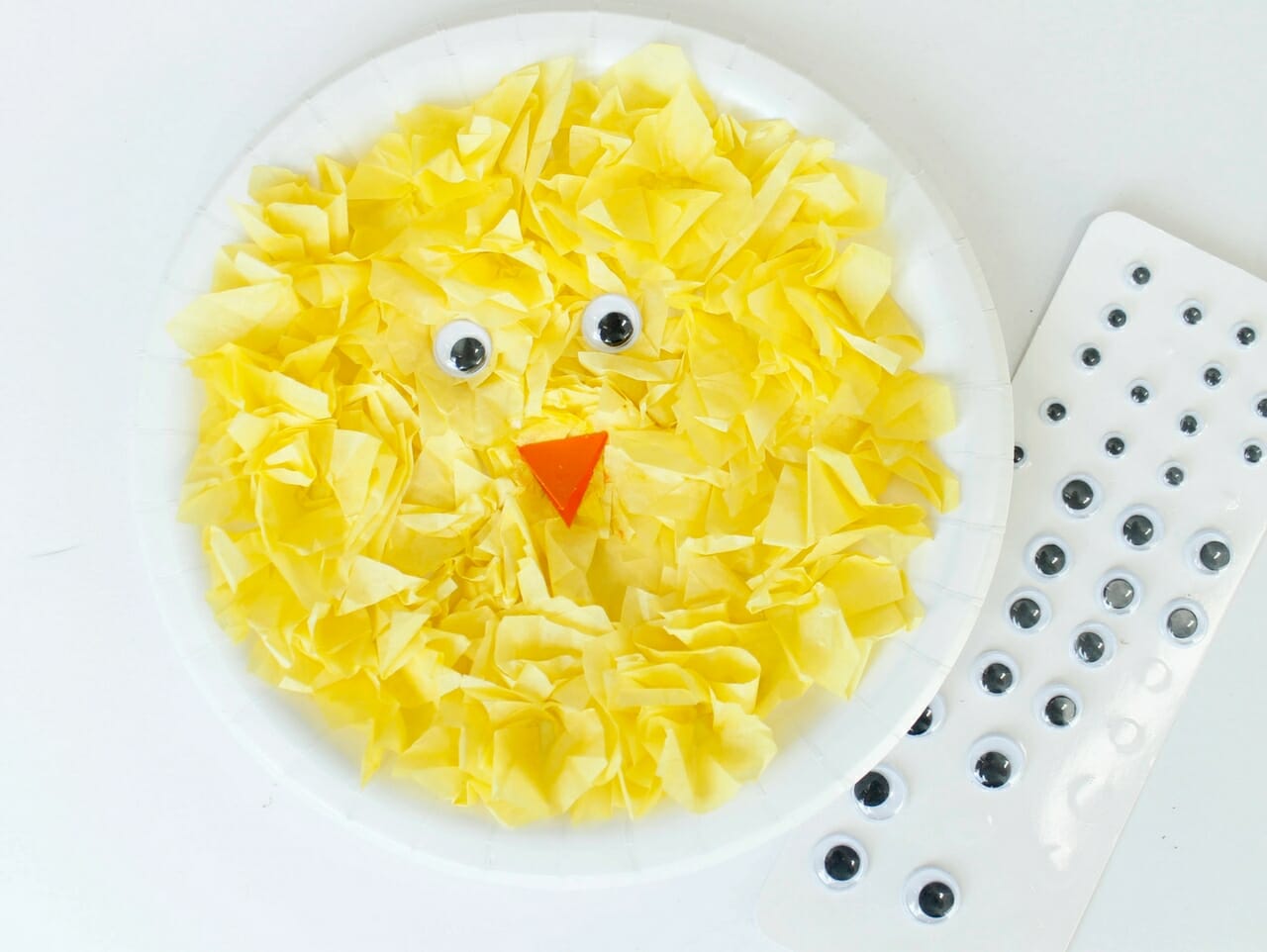 White background with white paper plate that has yellow tissue paper glued on to make a yellow chick and googly eyes on the side.